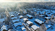 Aerial photo of a rural area filled with snow-covered detached houses, surrounded by snowy trees drenched in sunlight.