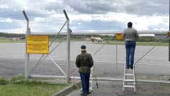 Ihmisiä seisomassa lentokentän vekkoaidan vieressä. Kuvaajilla on myös tikkaat, joilla he pääsevät aidan yläpuolelle.
