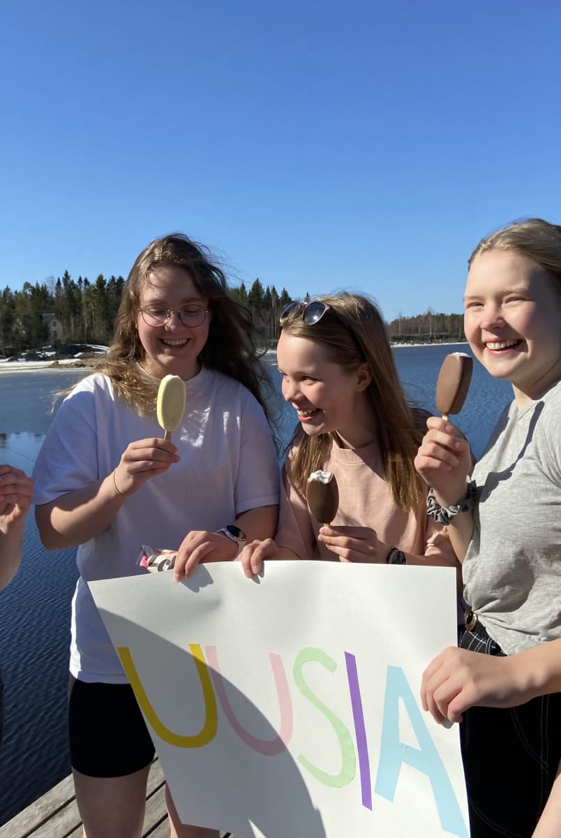 Kuvassa tytöt syövät jäätelöä laiturilla ja pitelevät kylttiä kädessään.