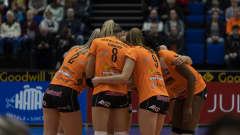 Oranssipaitaiset naislentopalloilijat seisovat lähekkäin ringissä.
