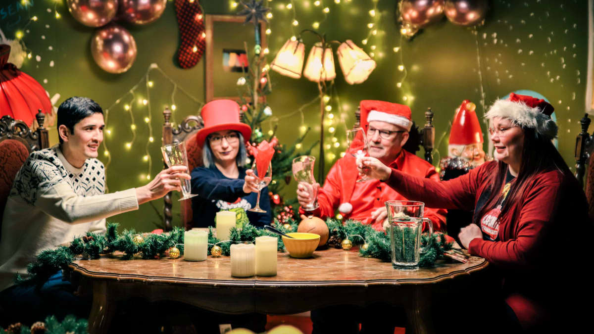 Värikkäisiin jouluasuihin pukeutuneet neljä ihmistä nostaa maljan jouluillallisella. Joulupöytä ja lavasteet ovat täynnä joulukoristeita.