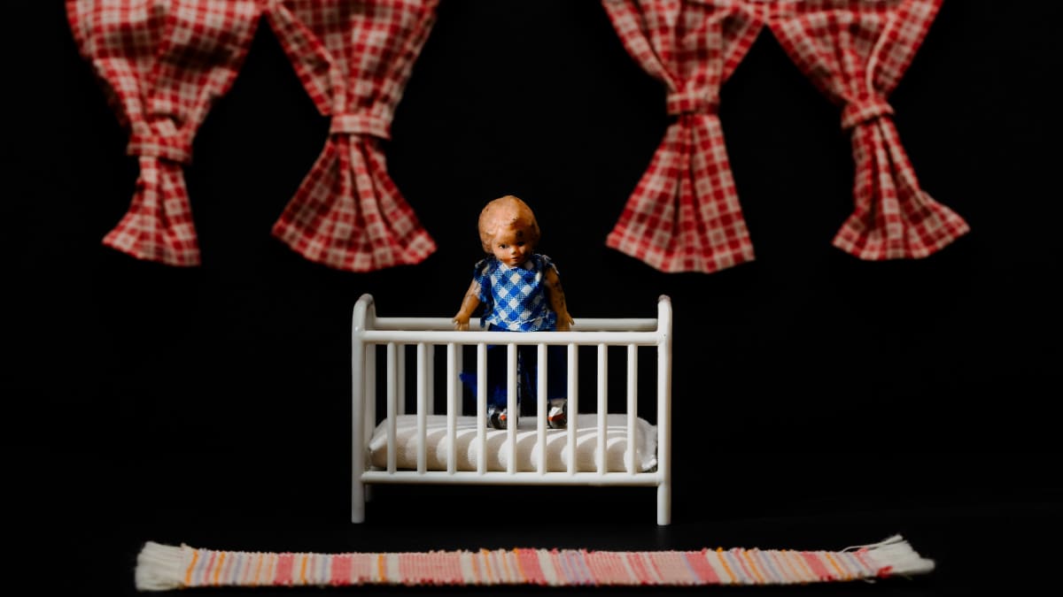 Miniatyyrinäkymässä pientä lasta esittävä nukke katsoo kameraan tuimasti pinnasängystä.