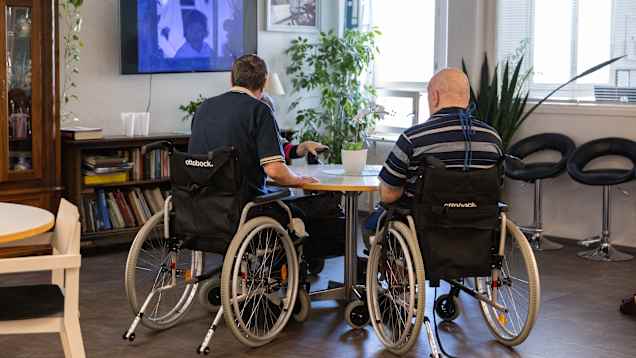 Kaksi henkilöä istumassa pyörätuoleissa, keskustelemassa olohuoneessa television ääressä