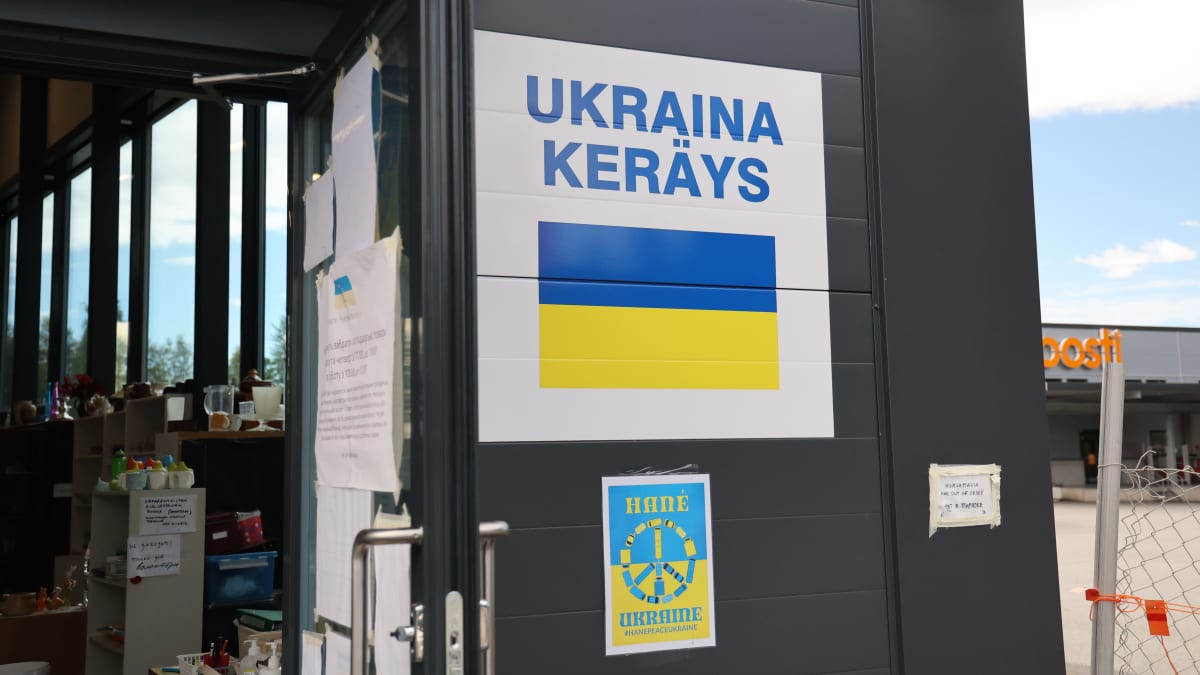 Suuri valkoinen kyltti harmaalla seinällä. Kyltissä lukee "Ukraina-keräys". Kyltin vieressä on avonainen ovi.