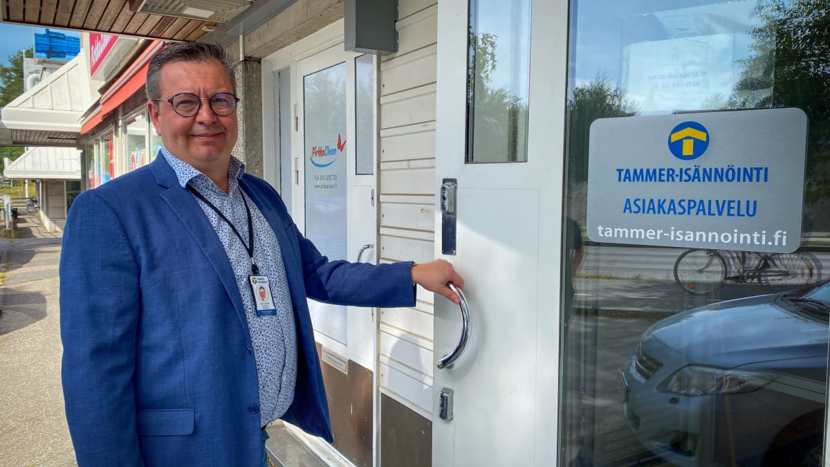 Mies sinisessä takissa ja kauluspaidassa pitää kiinni oven kahvasta ja hymyilee kameralle. Ovessa lukee "Tammer-Isännöinti asiakaspalvelu".