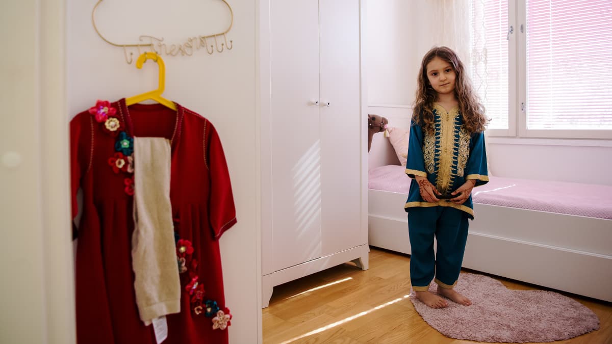 Liliya poseeraa huoneessaan, vasemmalla odottavat huomisen juhlapäivän vaatteet.