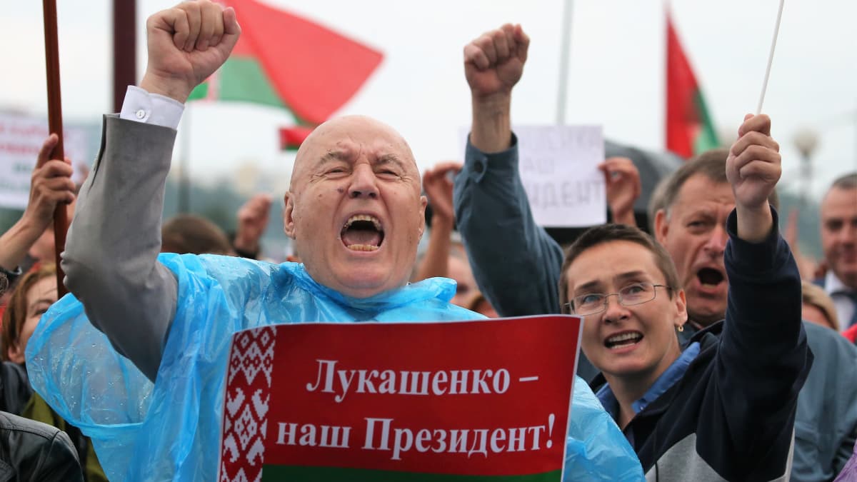 Mielenosoitusta Minskissä, Etualalla siniseen sadeviittaan pukeutunut mies pitää oikeaa kättään nyrkissä ja toisella Valko-Venäjän lipun väreillä koristeltua lappua, jossa lukee: Lukashenka - meidän presidenttimme!
