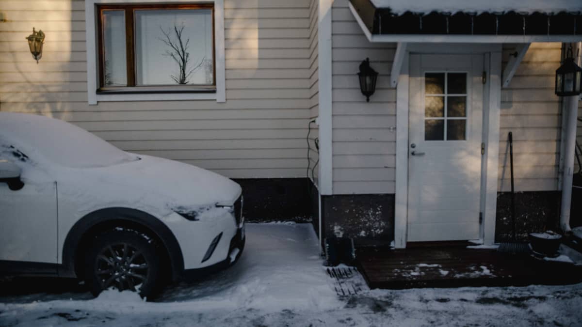 Valkoinen luminen auto seisoo valkoisen puutalon edessä.