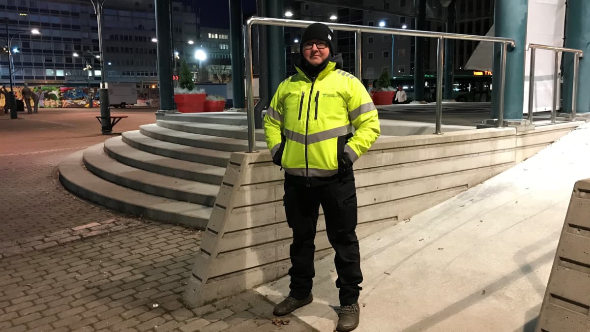 Mies seisoo tyhjällä torilla. Miehellä on yllään keltainen heijastintakki.