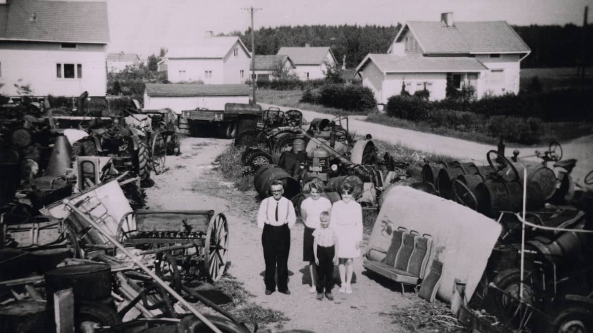 Nelihenkinen perhe seisoo romuvarastoa halkovalla kujalla. Taustalla rintamamiestaloja.