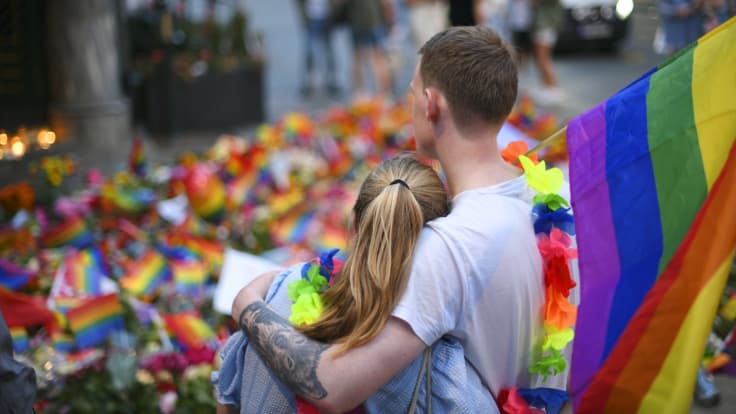 Kaksi henkilöä nojaa toisiinsa Pride-lippu olalla, taustalle on laskettu kukkia.