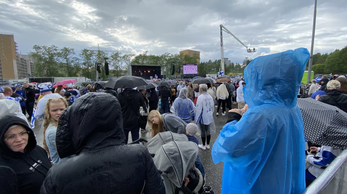 Ihmisiä sadetakeilla ja sateenvarjoila varustettuna odottamassa kansanjuhlan alkamista Hakametsän parkkialueella.