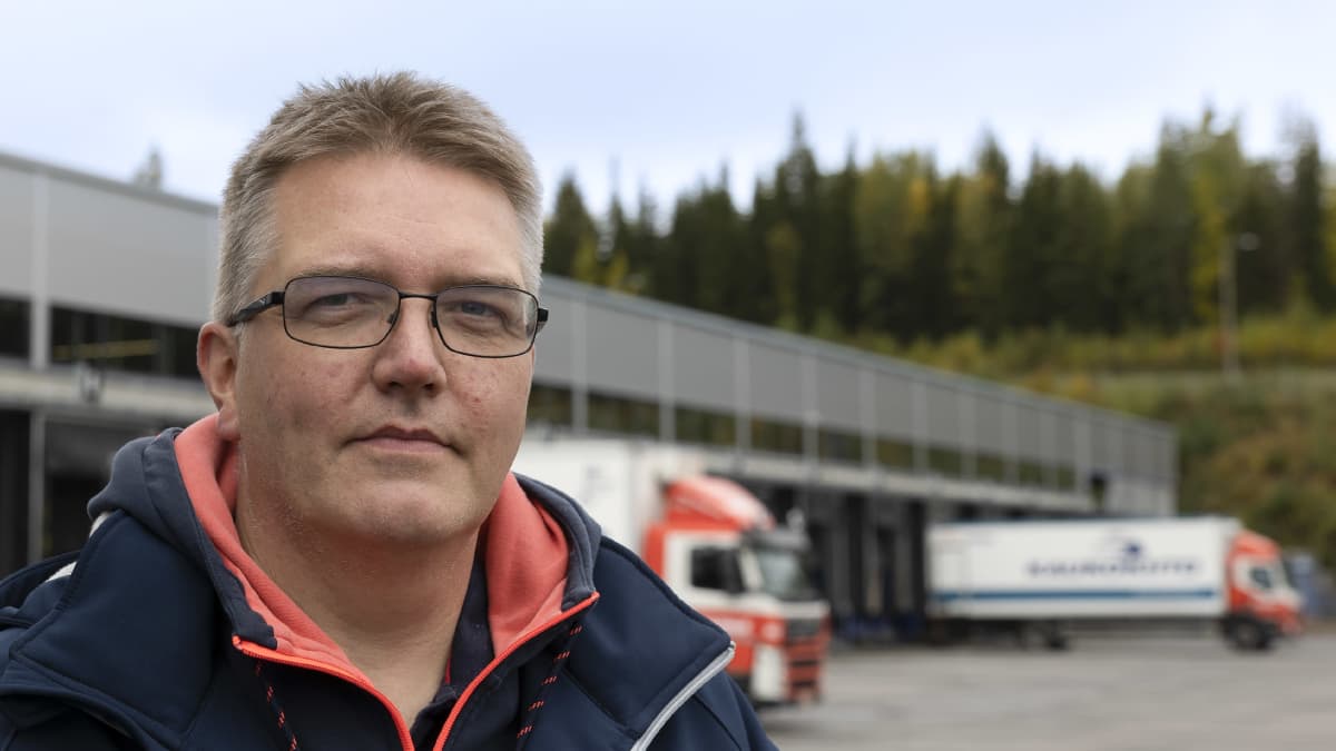 Kuljetusliike Taipaleen yrittäjä Riku Vainio katsoo kameraan Taipaleen autoterminaalin edustalla. Hänen takanaan siintää kaksi kuorma-autoa.