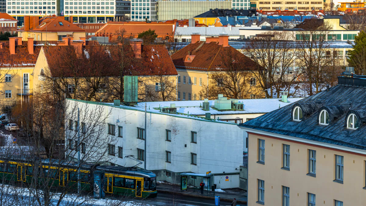 Tiivis kaupunkimaisema, talvinen aurinko paistaa kerrostalojen katoille, raitiovaunu on saapumassa pysäkille.