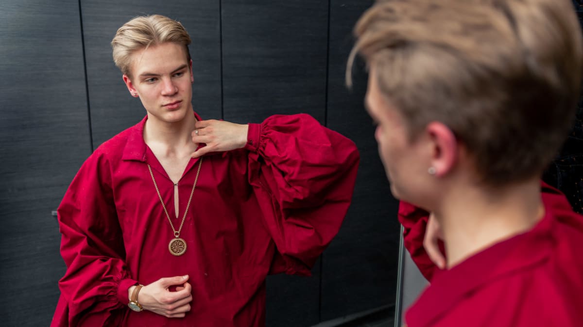 Jaakko Rikalainen peilaa itseään peilistä. Päällä hänellä on punainen isohihainen paita ja kalulassa pyöreä kalevala koru.