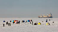 Ihmisiä ja kalastajien telttoja merijäällä, taustalla näkyy konttilaiva.