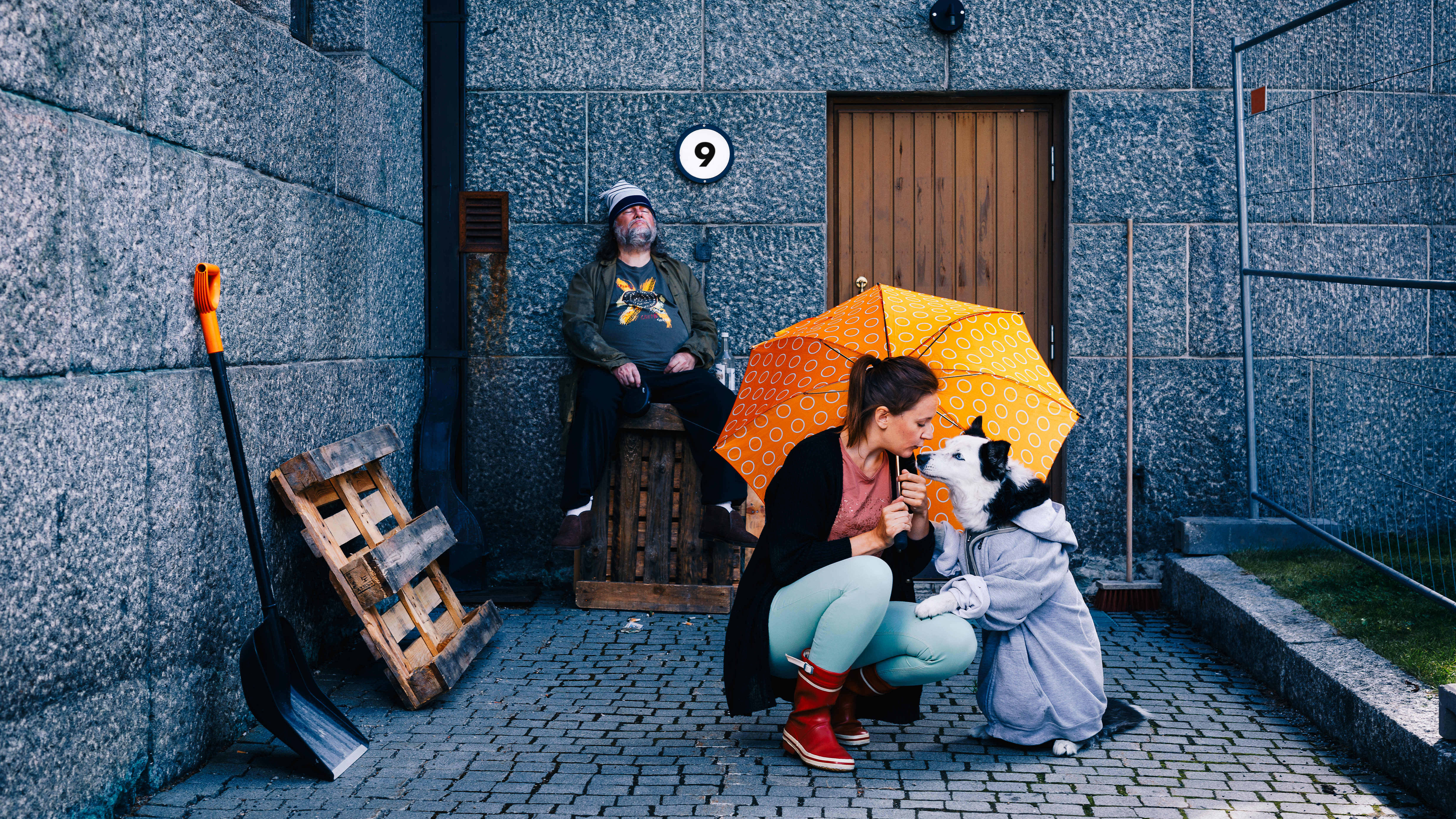 Nainen ja koira ovat oranssisen sateenvarjon alla. Partainen mies istuu taustalla seinän vieressä.