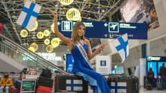 Essi Unkuri poseeraa lentokentällä kameralle sinisessä iltapuvussa, istuu matkalaukkujen päällä ja pitelee Suomen lippua.
