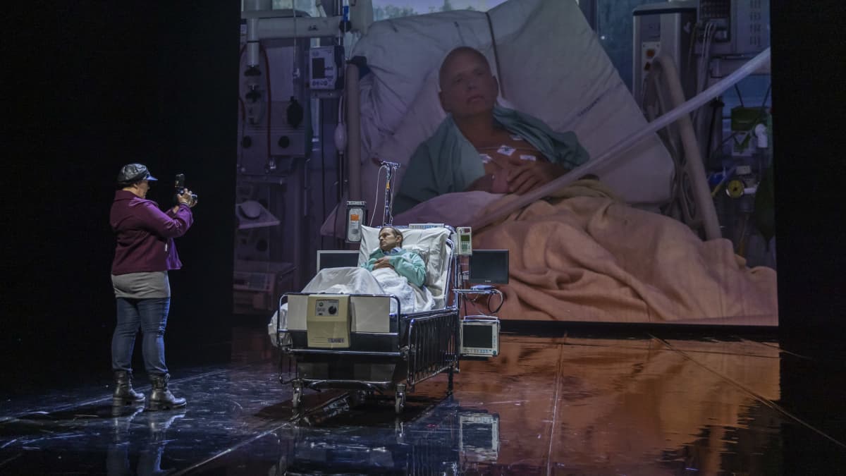 Nainen kuvaa sairaalasängyssä makaavaa miestä. Kuva näkyy taustaheijastuksessa.