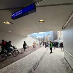 Cyclists and pedestrians in a tunnel with buildings, blue sky and bare trees in the distance.