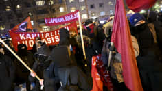 A crowd of people, mostly in black with their faces covered, some holding red flags and banners, in a city square on a cold night.