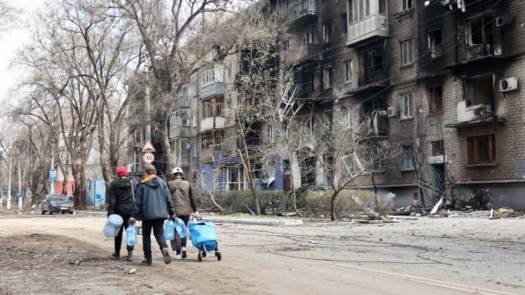 Ihmiset kantavat tyhjiä vesikanistereita tuhoutuneen kerrostalon vieressä.