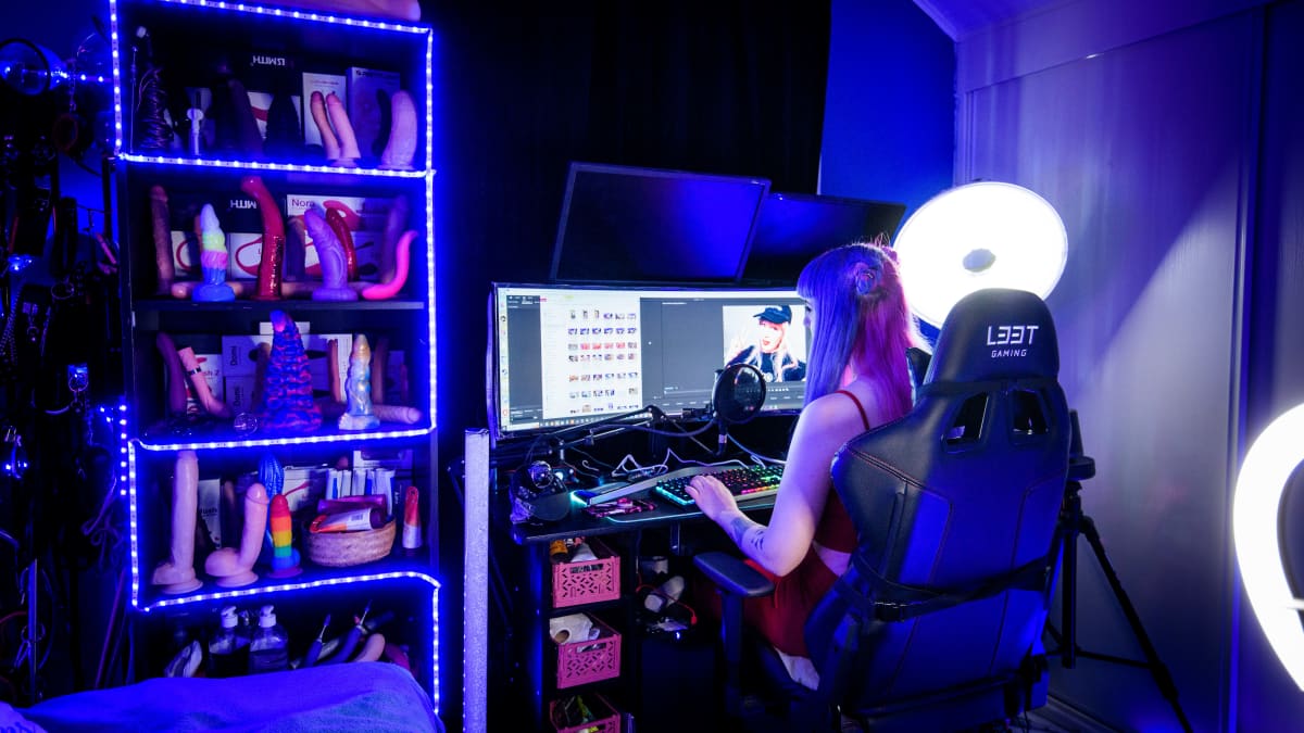 Nainen istuu tietokoneella huoneessa, jossa on studiovaloja ja kirjahylly täynnä seksileluja. 