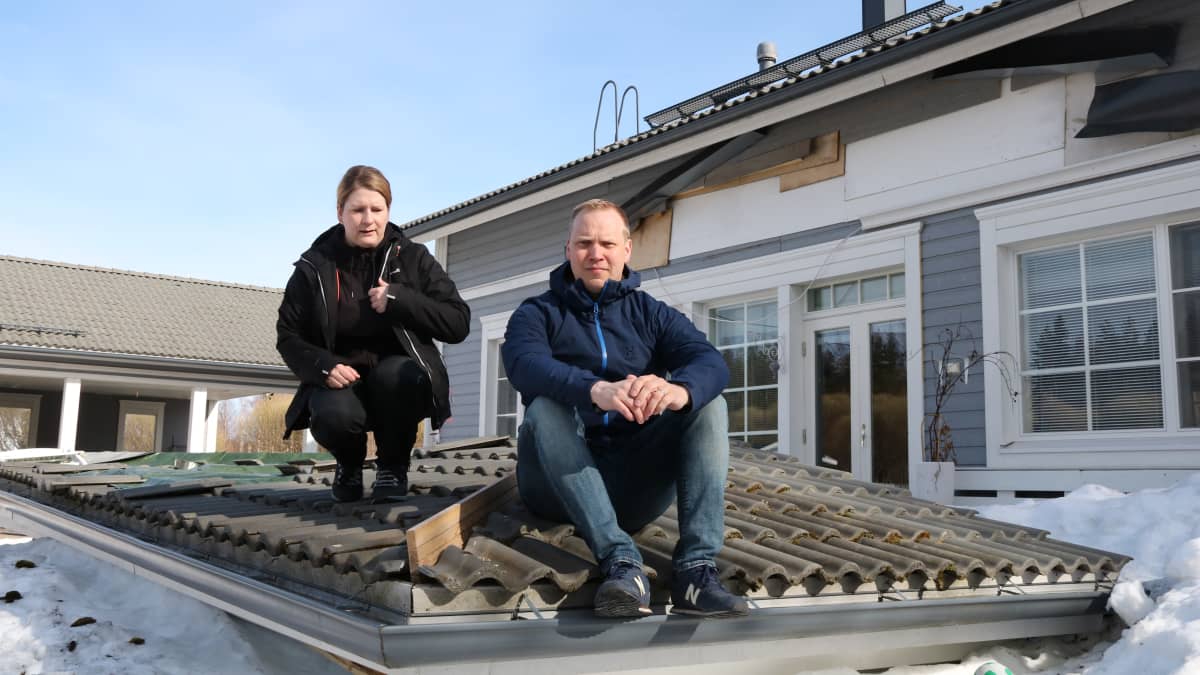 Mies ja nainen istuvat romahtaneen terassin katon päällä lumisella pihamaalla. Taustalla omakotitalo, jonka seinässä on reikä.