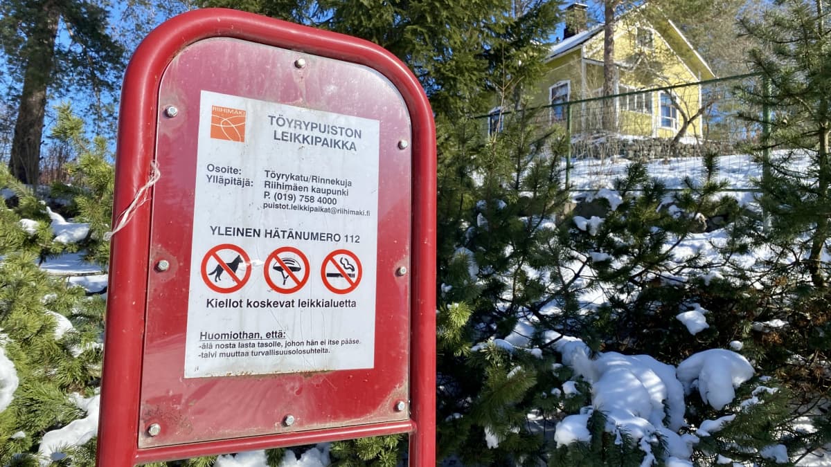 Punainen kyltti leikkipuistossa. Kyltissä kerrotaan yleinen hätänumero ja leikkialueen säännöt.