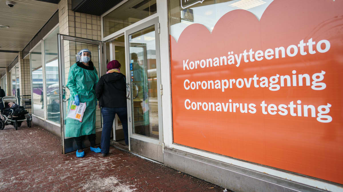 Henkilö menossa sisään koronatestauspaikkaan. Henkilökunnan edustaja pitää ovea auki. Ikkunassa teksti koronanäytteenotto kolmella kielellä.