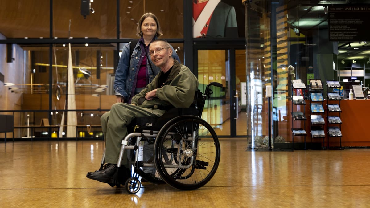 Juha Hinkkanen istuu pyörätuolissa ja hymyilee aulatilassa. Takana seisoo Leila Hinkkanen.