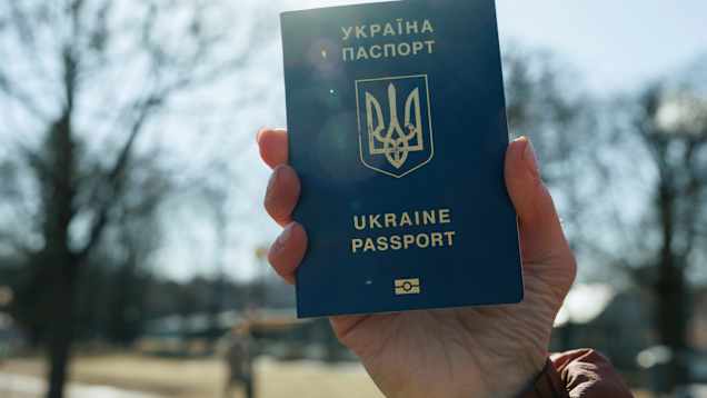 Naisen käsi pitelee Ukrainan passia. Taustalla taivasta ja lehdettömiä puita.