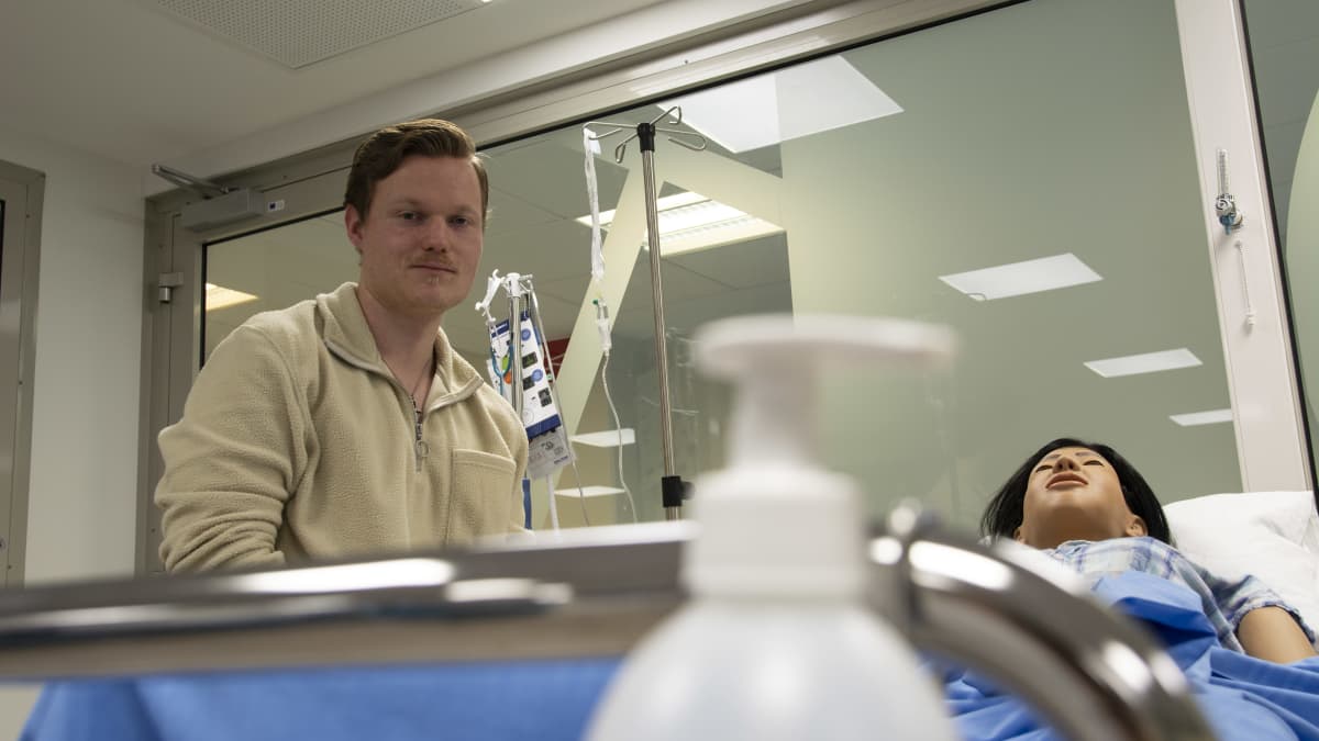 Sairaanhoitajaopiskelija Jonne Häyrynen istuu potilasvuoteen vieressä ja katsoo suoraan kameraan.