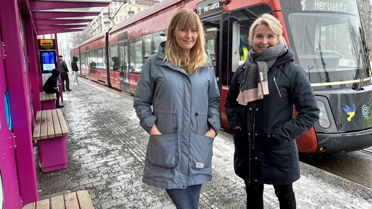 Toimittajat Anna Sirén ja Katri Tapola seisovat ratikkapysäkillä, taustalla näkyy ratikka.