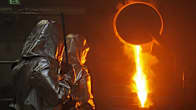Two people in silver coloured heat protection gear next to a bright orange molten gold being poured out of a blazing hot furnace.