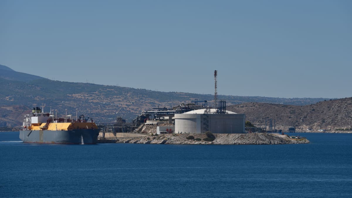 Mereltä päin kuvattuna satama, jossa vasemmalla näkyy sininen, ruosteinen iso LNG-alus ja keskellä kaasusäiliö. Taustalla kuivaa kreikkalaista maisemaa.