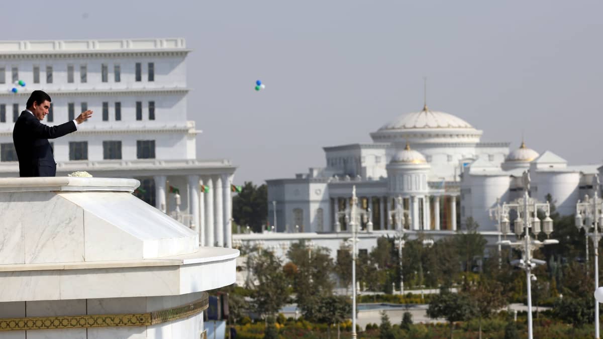 Turkmenistanin presidentti seisoo palatsin parvekkeella mustassa puvussa ja vilkuttaa alaspäin. Taustalla näkyy suuria valkoisia palatsimaisia rakennuksia.