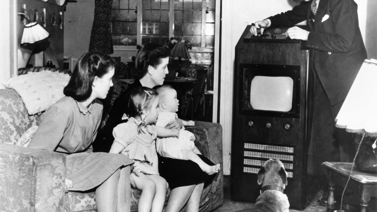 Mustavalkokuvassa perhe katsoo televisiota. Äiti vauva sylissään ja kaksi tytärtä istuu sohvalla, koira makaa lattialla ja perheenisä säätää televisiovastaanotinta.