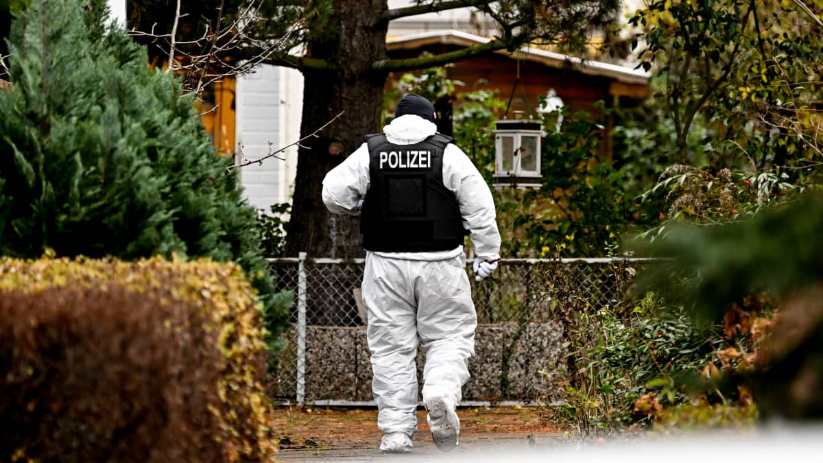 En polis i vit skyddsdräkt och mörk skyddsväst med texten Polizei på ryggen går mellan häckar i ett småhusområde i höstfärger.