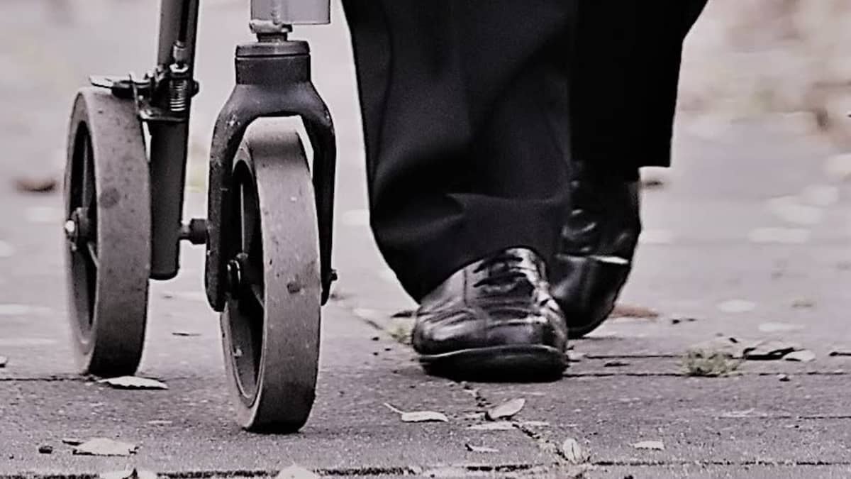 Någon går med en rollator, svarta skor och byxor och rollatorns hjul och nedre del syns. Nästan svartvit bild. 