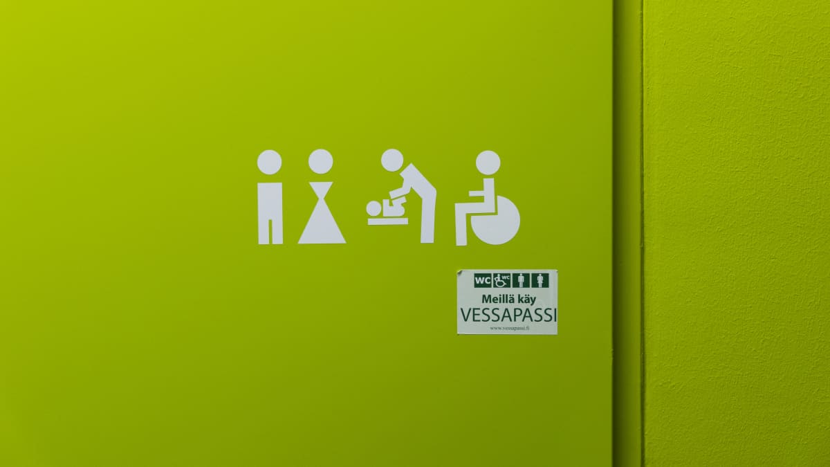 Vihreällä taustalla valkoisia symboleita: Mies, nainen, lastenhoito, pyörätuoli. Niiden alla tarra, jossa lukee "Meillä käy vessapassi".
