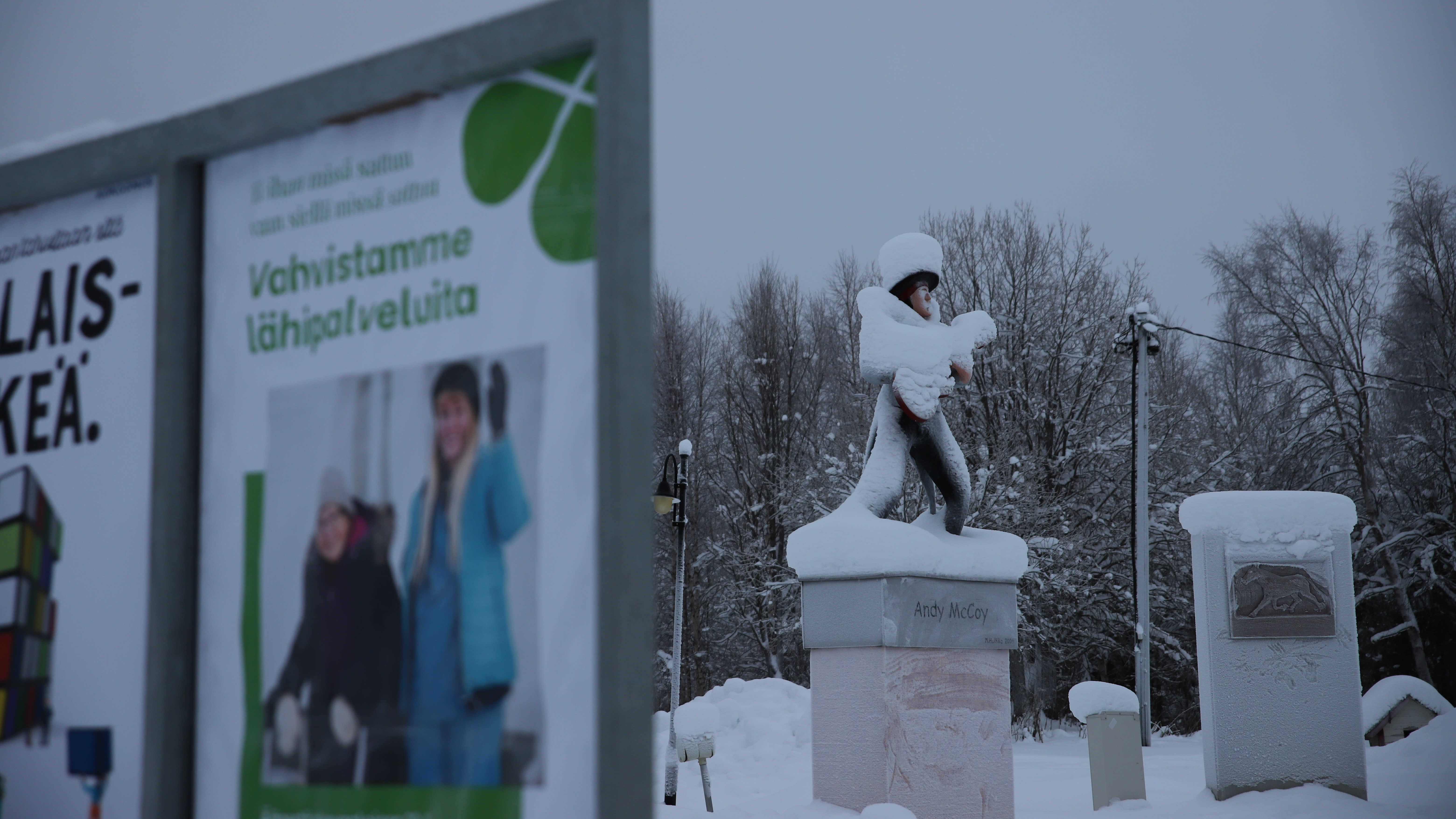 lumen peittämä Andy Mc Coyn patsas ja vaalimainos Pelkosenniemen kirkonkylällä