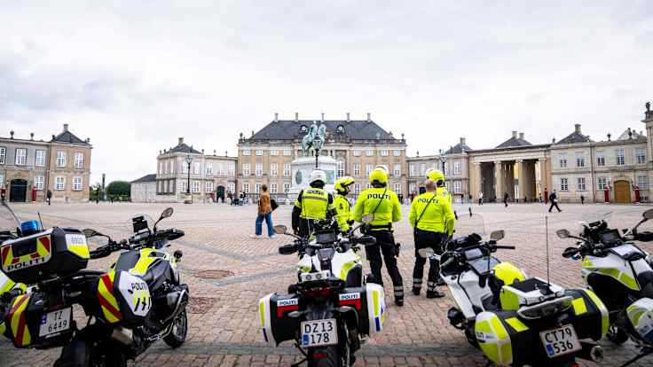 Four people wearing yellow jackets with the word POLITI stand near four motorcycles on a square surrounded by ornate old buildings.