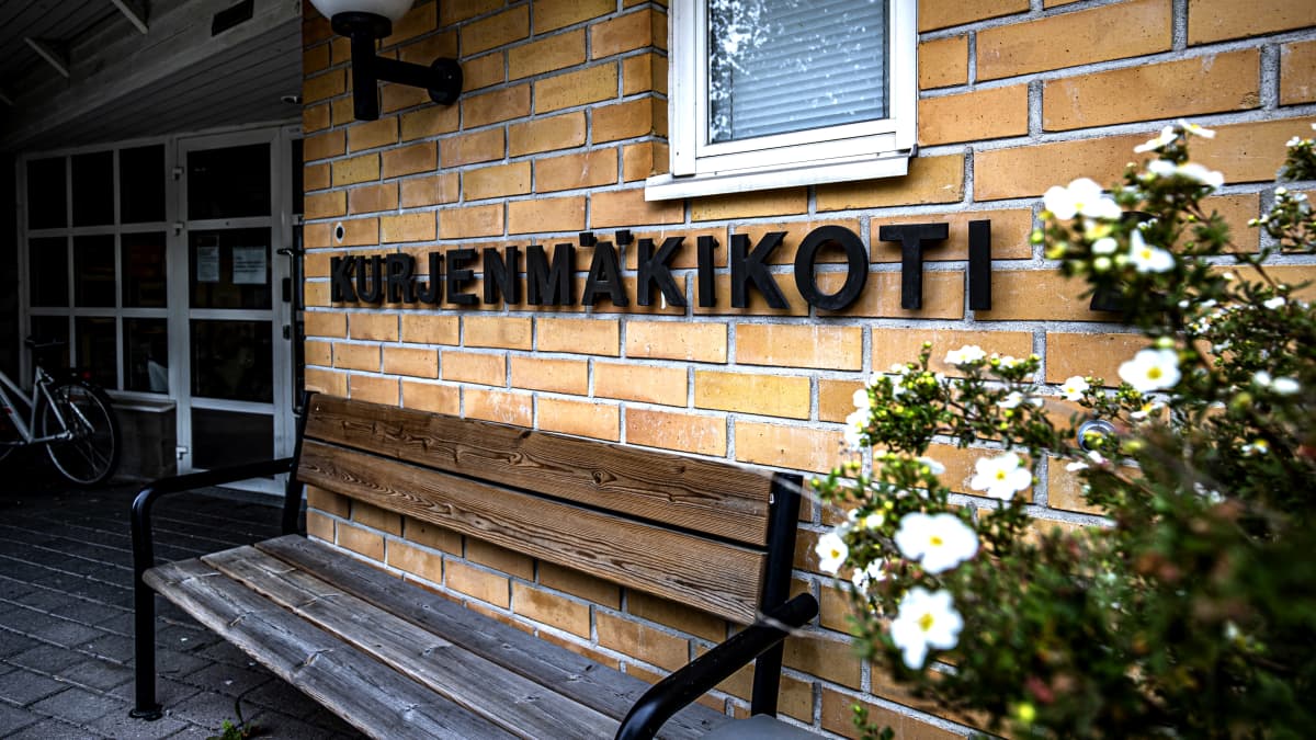 En gul tegelvägg med texten kurjenmäkikoti 2. Framför väggen finns en sliten bänk.