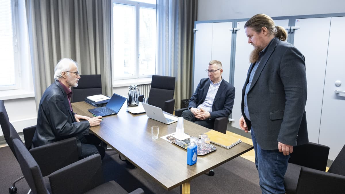 Tre män kring ett bord på riksförlikningsmannens byrå: till vänster sitter förlikningsman Leo Suomaa, i mitten sitter AKT:s ordförande Ismo Kokko och till höger står arbetsmarknadschef Juha Mutru.