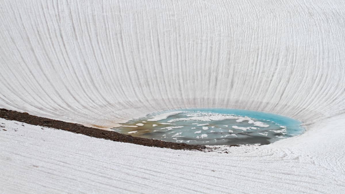 Sulava lumi muodostaa pienen järven, jossa kelluu jäälauttoja. Sulaneen lumen veden virtaus näkyy rantuina lumella.