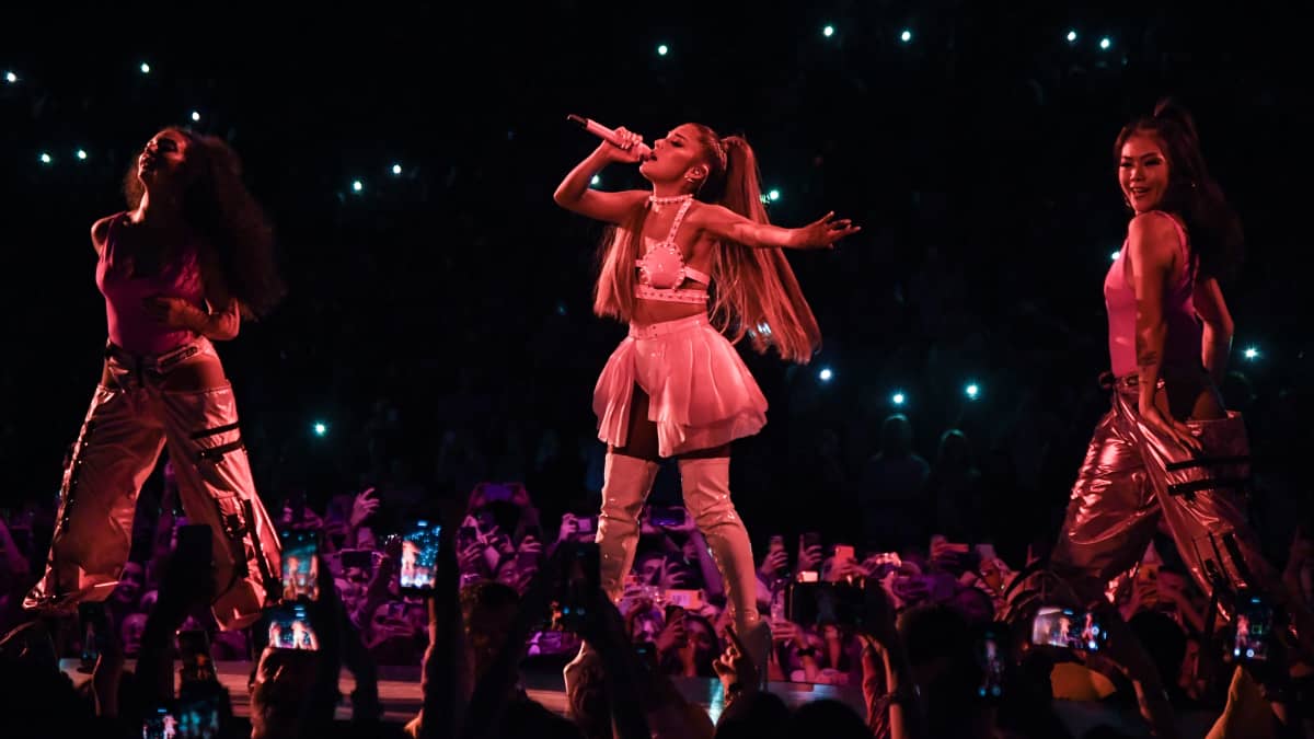 Laulaja Ariana Grande laulaa silmät kiinni lavalla kahden tanssijan ympäröimänä. Hänellä on yllään valkoinen latex-toppi, lyhyt vaalea röyhelöminihame ja korkeat valkoiset latex-saappaat.