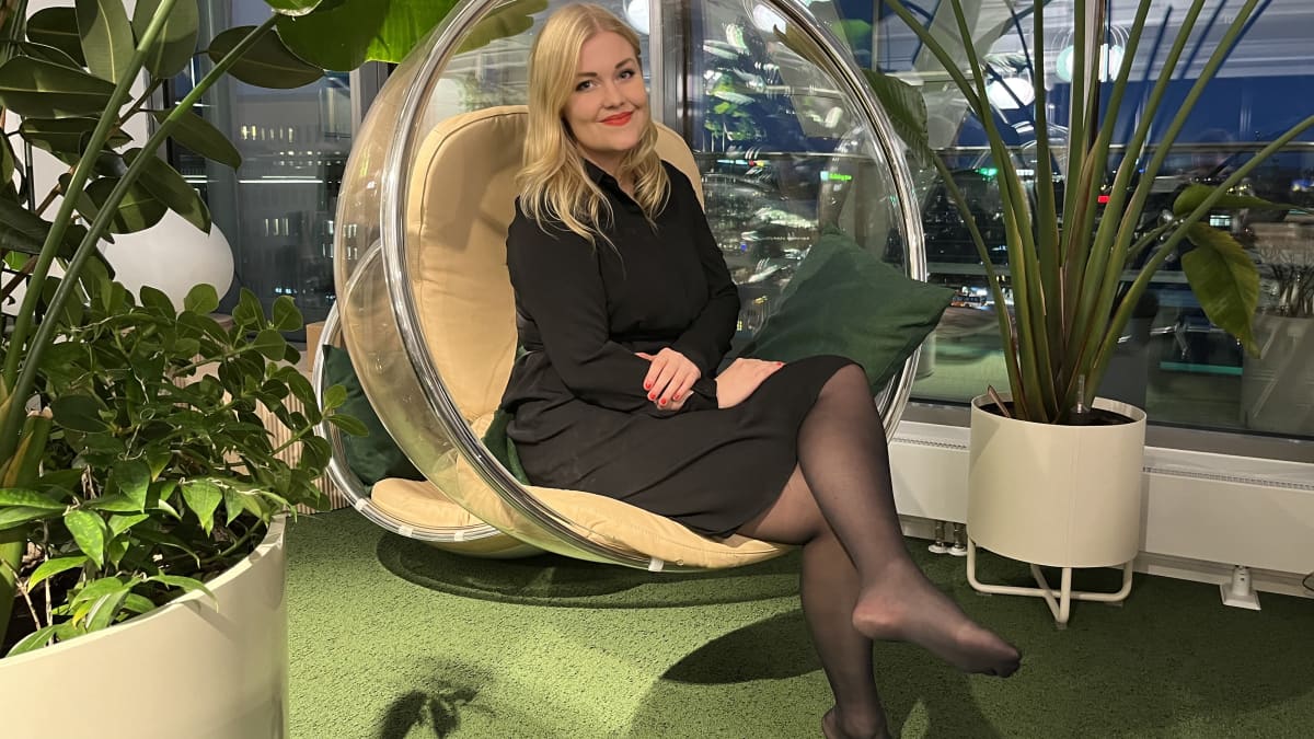Laura "lapa" Kyntäjä istuu pehmustetussa ja läpinäkyvässä pallokiikussa, ympärillä viherkasveja ja ikkunasta näkymät päärautatieasemalle Helsingissä
