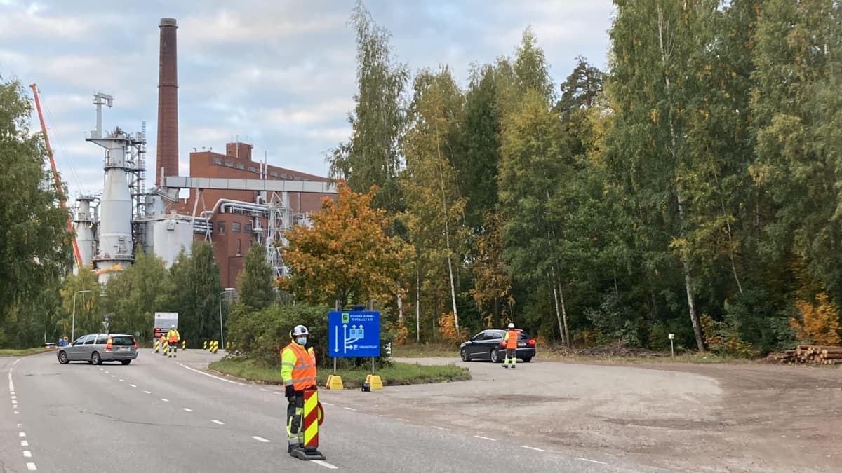 Neonvärisiin työvaatteisiin pukeutunut mies ohjaa autoja tiellä, taustalla mestää ja tehdas.
