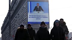 Ihmiset kävelevät kadulla Moskovassa, kun heidän takanaan olevalla näytöllä näkyy Vladimir Putin pitämässä vuotuista puhettaan liittokokoukselle.